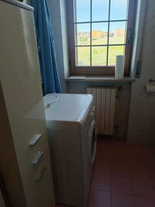 Laundry room with washing machine, window overlooking Atessa cityscape, radiator, and blue curtains in Tales of Abruzzo