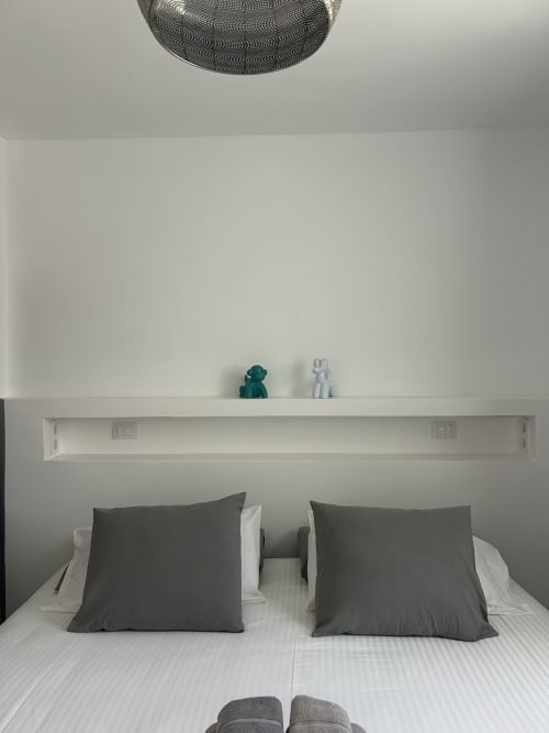 Moderno dormitorio con almohadas grises, ropa de cama blanca, ventilador de techo y estantería decorada en Kamalahouse SL, Corralejo