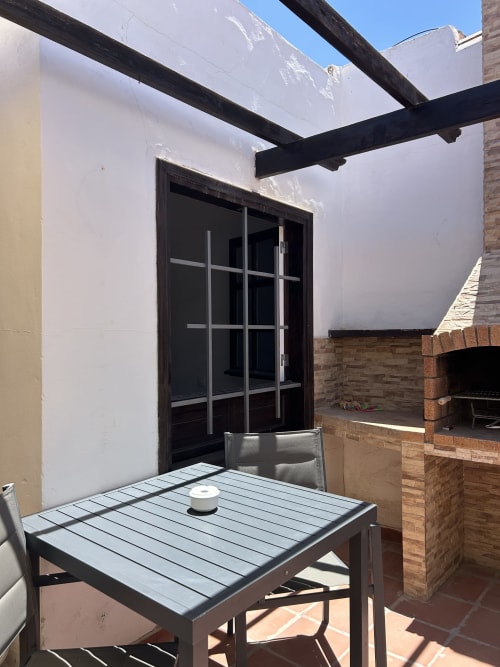 Terraza soleada en Kamalahouse SL en Corralejo con mesa de comedor, pérgola negra, paredes blancas y zona de barbacoa de ladrillo