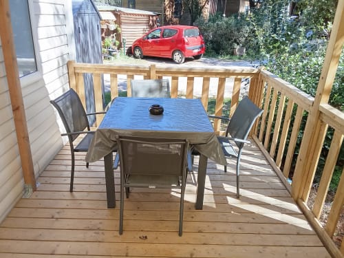 Terrasse en bois avec table de salle à manger et chaises au mobil-home Camping La Lézardière, vue sur le jardin avec voiture rouge garée à proximité