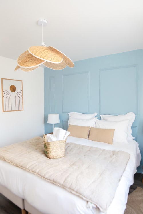 Chambre lumineuse de la villa ARLOÉ avec tête de lit en panneaux bleus, suspension en rotin, literie blanche et décoration côtière
