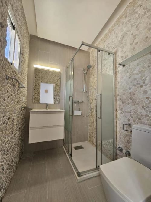 Baño moderno en el hostal Villa Palmer de Paguera con ducha a ras de suelo, lavamanos blanco, inodoro y paredes de mármol