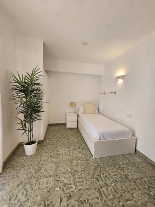 Moderno dormitorio individual en el hostal Villa Palmer de Paguera con cama blanca, planta decorativa, suelo con patrón y aplique de pared