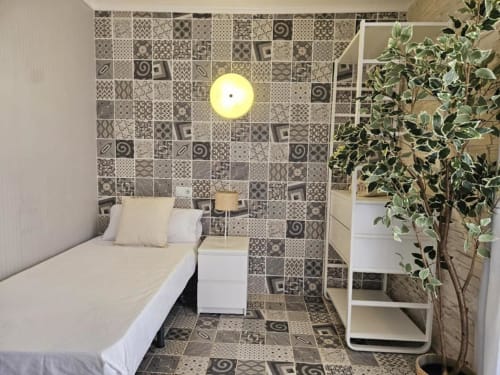 Habitación individual compacta en Villa Palmer con paredes de azulejos estampados, cama sencilla, mesita blanca, planta en maceta y mesa redonda