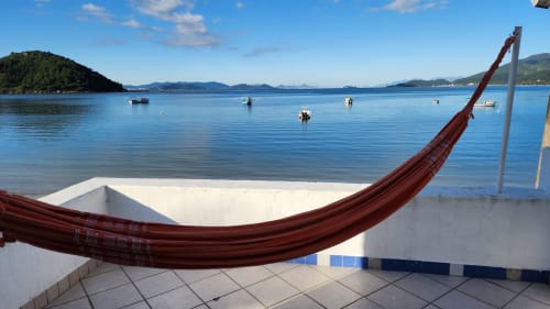 Rede vermelha no terraço à beira-mar do hotel Philia em Florianópolis, com vista para a baía azul tranquila e barcos ancorados
