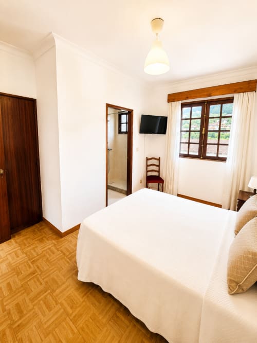 Quarto duplo luminoso na Pousada Serrana Gerês com cama branca, móveis de madeira, TV e vista para as montanhas através de amplas janelas