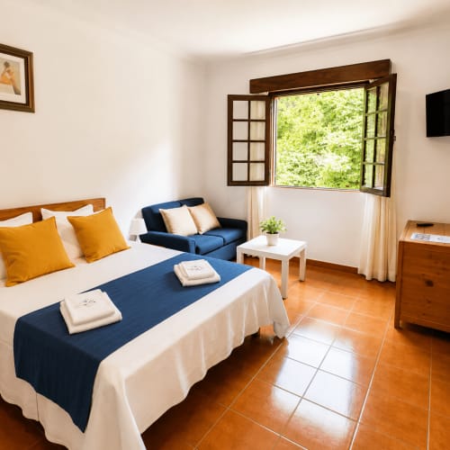 Quarto luminoso na Serrana Gerês Inn com cama azul, almofadas amarelas, sofá e piso em terracota com vista para as montanhas
