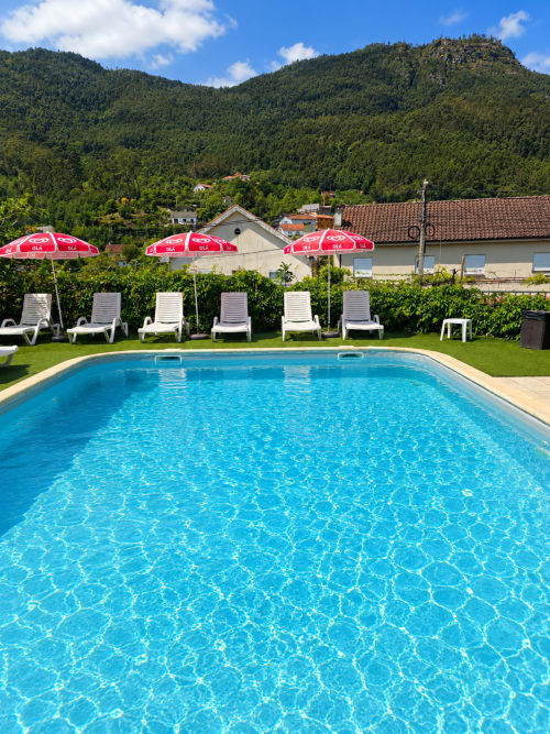 Piscina reluzente da Pousada Serrana Gerês com espreguiçadeiras brancas, guarda-sóis vermelhos e montanhas arborizadas ao fundo