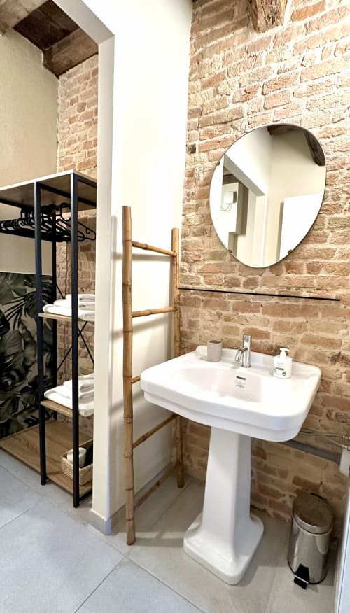 Bagno moderno nell'appartamento NOUA LIVING Ravenna con muri in laterizio faccia a vista, lavandino a piedistallo, specchio rotondo e asciugamani in legno