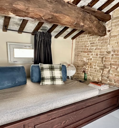 Accogliente camera con loft a NOUA LIVING Ravenna: travi in legno a vista, muri in mattoni, cuscini blu, cuscino scozzese e lampada.