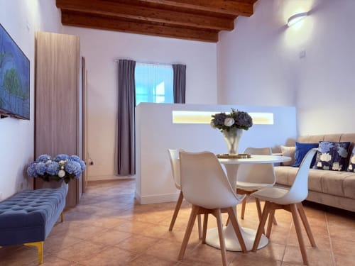 Moderno appartamento a Cagliari con zona pranzo, sedie bianche, tavolo rotondo, divano beige, fiori blu e travi in legno