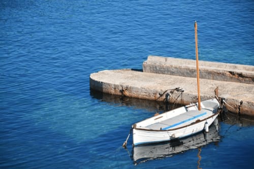 Barco de pesca blanco y azul amarrado en el muelle de Es Canar con aguas azules cristalinas del Mediterráneo, cerca de Apartamentos Playa Sol
