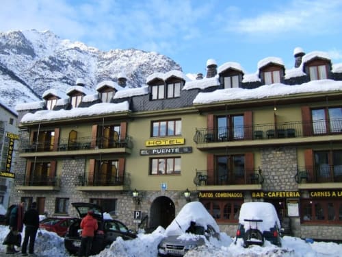 Hotel El Puente en Benasque con tejado nevado, Pirineos de fondo y coches aparcados