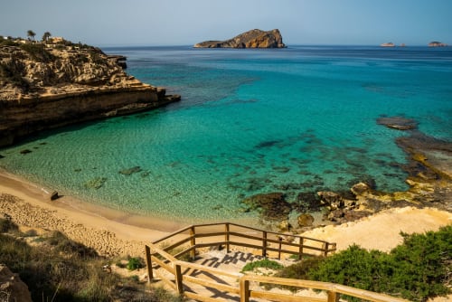 Escaleras de madera para acceso a la playa con vistas a aguas turquesas cristalinas y arena blanca en Es Canar, Ibiza