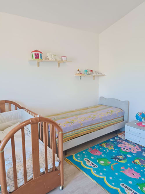 Bright children's bedroom with wooden crib, toddler bed, colorful play mat, and wooden shelves with toys in Abruzzo apartment
