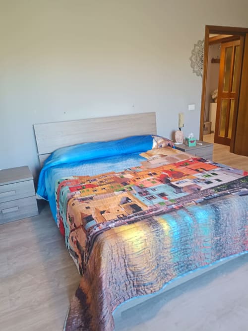Bright bedroom with queen bed featuring colorful cityscape bedding, blue pillows, wooden headboard, nightstand, and wooden