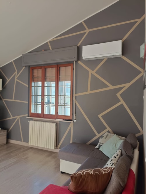 Modern apartment bedroom with geometric wall pattern, red-framed window, split AC units, radiator, and sofa at Tales of