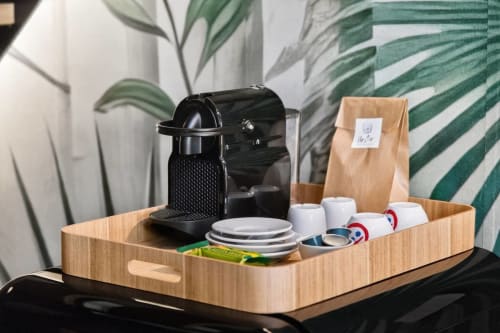 Vassoio di benvenuto nell'appartamento Nestco Milano con macchina da espresso, tazze, piatti e caffè in scatola di legno su sfondo di foglie tropicali
