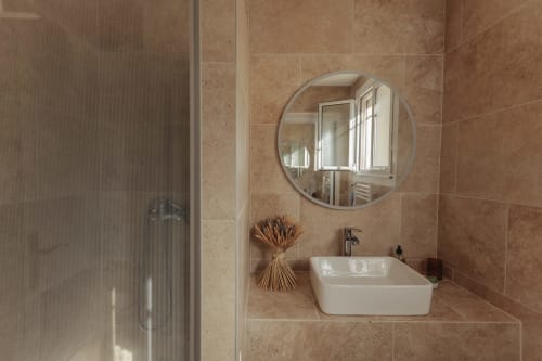 Salle de bain moderne du gîte Mas Tihou avec miroir rond, vasque blanche et murs en carrelage beige surplombant le jardin