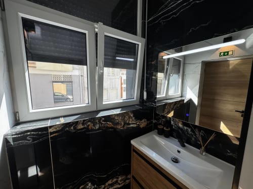 Banheiro moderno com paredes em mármore preto, pia branca, espelho LED e móvel em madeira na Vila Amber Star Rent, Vila Nova de Gaia