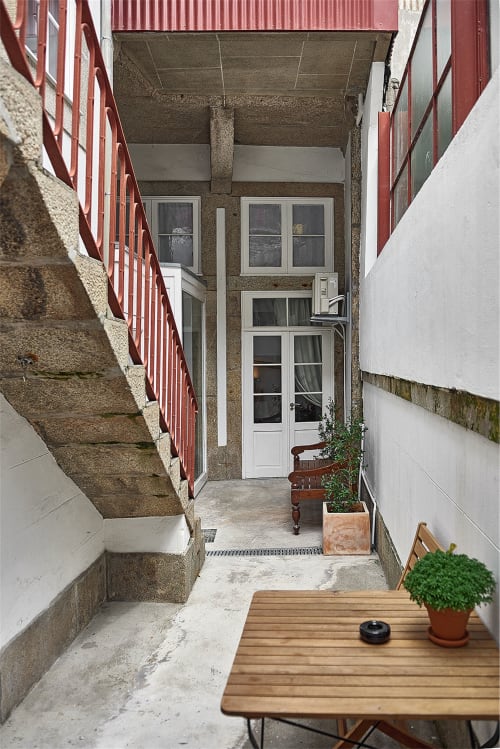 Entrada de pátio exterior estreito no PortoSense Almada no Porto, com escadas vermelhas, porta branca, plantas em vasos e mesa de madeira