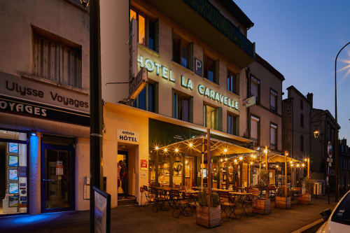 Hotel La Caravelle exterior at dusk in Riom, France with lit terrace seating and glowing signage
