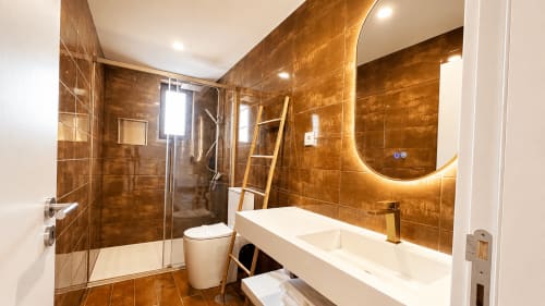 Banheiro de luxo moderno na Quinta das Susandas com azulejos bronze, espelho iluminado curvo, chuveiro aberto e branco