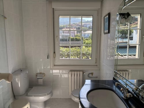 Baño luminoso en casa de Baiona con vistas al mar a través de ventana de cristales, azulejos blancos, inodoro, lavabo y radiador.