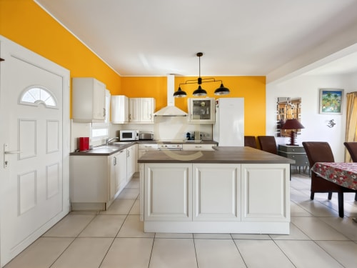 Cuisine moderne aux armoires blanches, mur d'accent jaune, îlot central et espace repas ouvrant sur le salon
