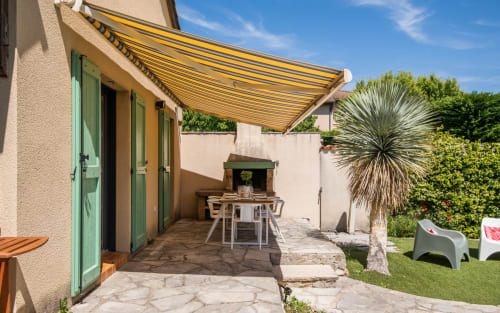 Villa patio with yellow awning, green doors, outdoor dining table, and palm tree in Millau