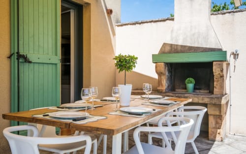 Outdoor dining patio with wooden table, white chairs, place settings, and green fireplace at Millau villa