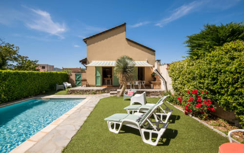 Millau villa with private pool, white lounge chairs on green lawn, red flowers, and beige house with green shutters