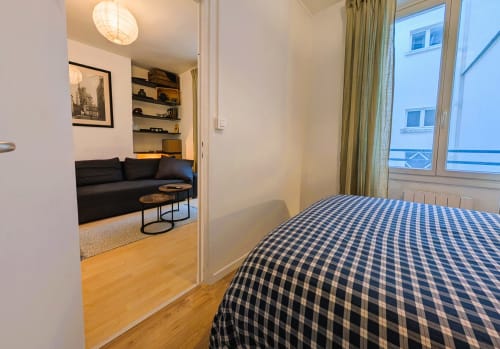 Parisian apartment bedroom with blue checkered bedding, wooden floor, and view into living room with black sofa and shelving