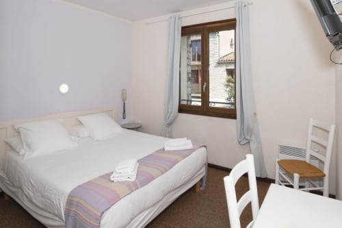 Chambre Lavande de l'Hôtel Le Verdon avec vue sur le village, literie blanche, chemin de lit violet, fenêtres brunes, équipements modernes