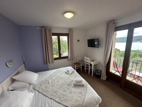 Chambre d'hôtel lumineuse avec mur d'accent violet, vue sur le lac depuis le balcon, volets en bois et mobilier moderne