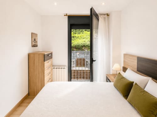 Dormitorio luminoso en apartamento Holibai de Baiona con vistas a piscina, muebles de madera, cojines oliva y puerta a terraza