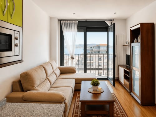 Luminoso salón moderno en apartamento de Baiona con sofá beige, mesa de madera y balcón con vistas al mar y cortinas.