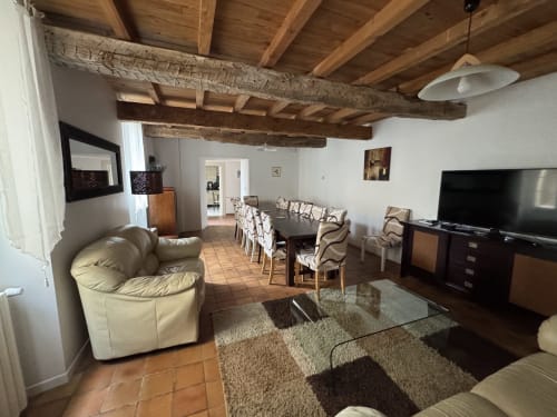 Spacieux salon de chalet avec poutres en bois apparentes, sièges en cuir crème, table à manger, TV et carrelage en terre cuite