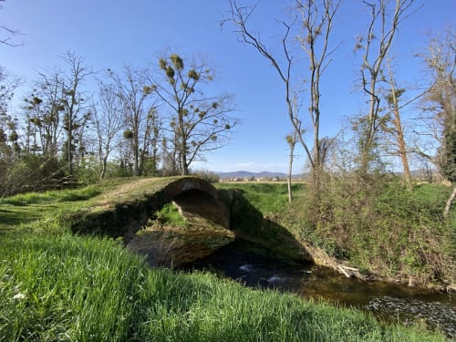 Pont en pierre cintrée enjambant un ruisseau avec arbres nus et prairies vertes en Ariège, près du gîte de Verniolle