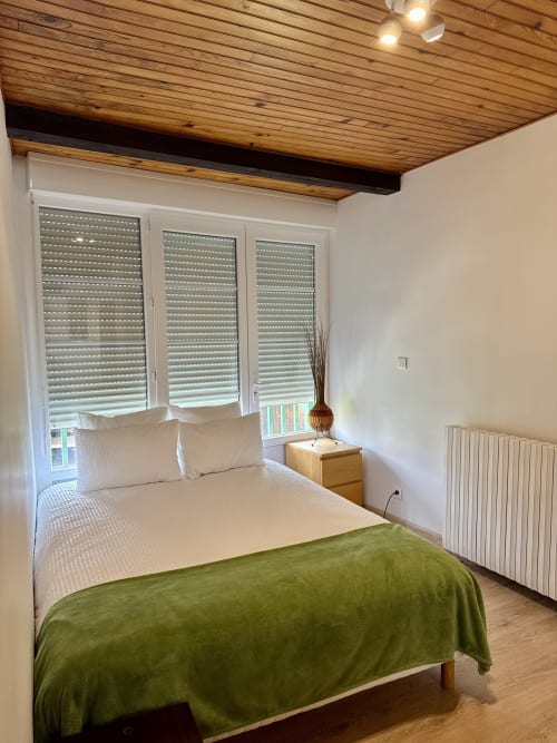 Chambre lumineuse du Gîte du Mercadal avec lit queen-size, plaid vert, literie blanche, plafond en bois et fenêtres avec volets