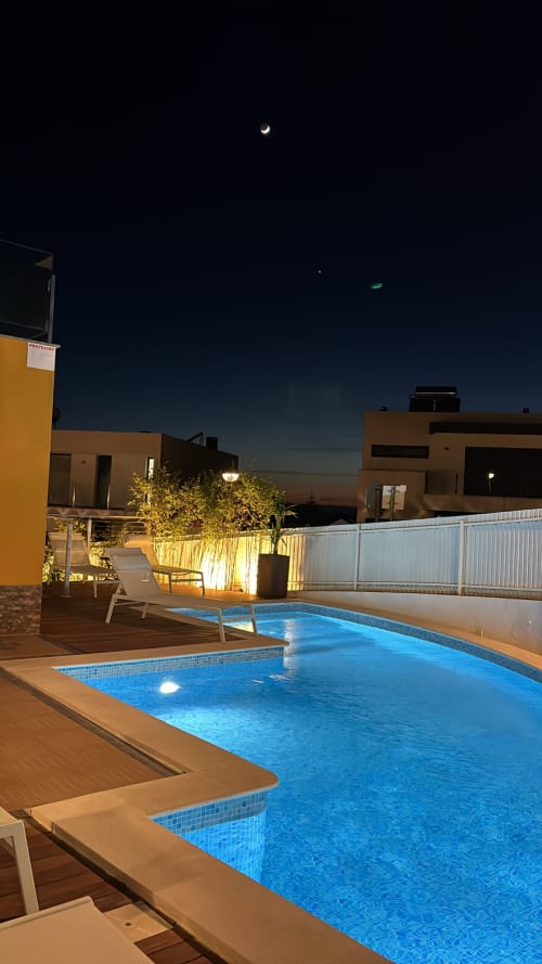 Piscina privada iluminada à noite com espreguiçadeiras, villa moderna, lua crescente e cerca branca em Sesimbra Sun