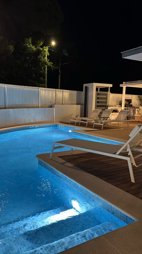 Piscina de resort iluminada à noite com espreguiçadeiras, grades brancas e estruturas de vilas em Sesimbra