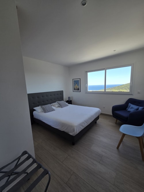 Chambre spacieuse avec vue sur mer à la Résidence Lésia, Corbara. Lit queen-size, fauteuil bleu, parquet, baies vitrées panoramiques