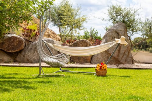 Cream hammock with straw hat in lush garden at Agriturismo Chiai Francesco, Bari Sardo, with wildflowers and rustic stones