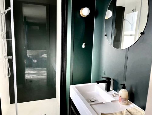 Salle de bain moderne du Domaine Pero Longo aux murs vert foncé, lavabo blanc, robinetterie noire, miroir rond et douche à effet pluie