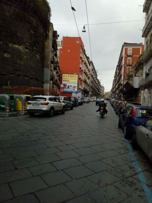 Bustling Naples street with parked cars, motorcyclist, and colorful residential buildings near Neapolis Tecnotourist