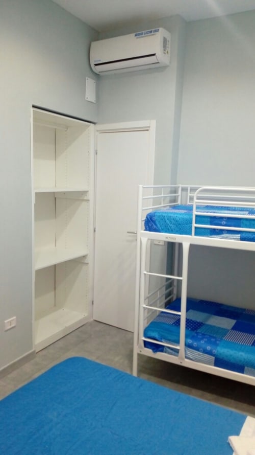 Bright bedroom with white bunk bed, blue bedding, open wardrobe, air conditioning unit, and blue floor mat at Neapolis