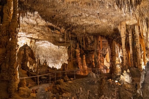 Cámara subterránea de cueva con estalactitas y estalagmitas iluminadas en tonos dorados, pasarela metálica visible, Villa Palmer Paguera