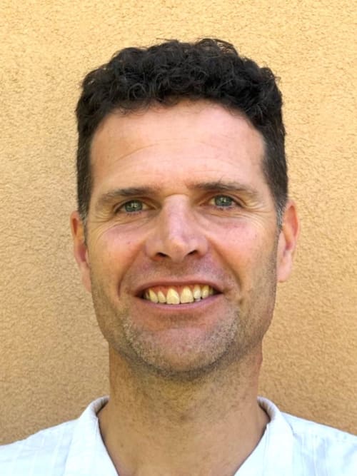 Homme souriant aux cheveux noirs et yeux verts devant un mur jaune au gîte Le Bacalan de Mormoiron