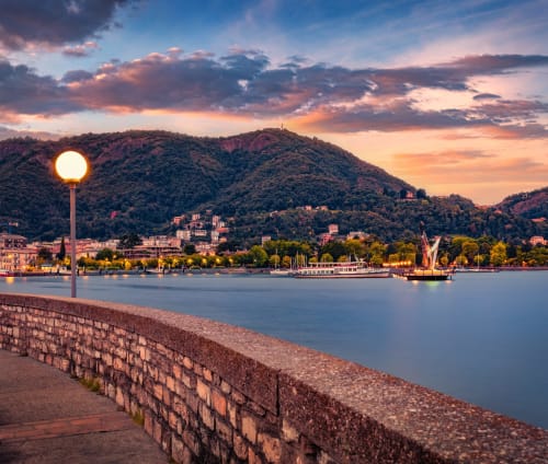 Vista del tramonto sul lungolago di Monza con lampione illuminato, barca a vela e colline boscose al crepuscolo